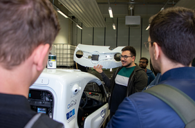 Ömer Dönmez zeigt einer Gruppe etwas im Labor vor einem autonomen Fahrzeug