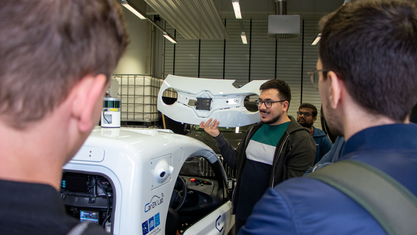 Ömer Dönmez zeigt einer Gruppe etwas im Labor vor einem autonomen Fahrzeug