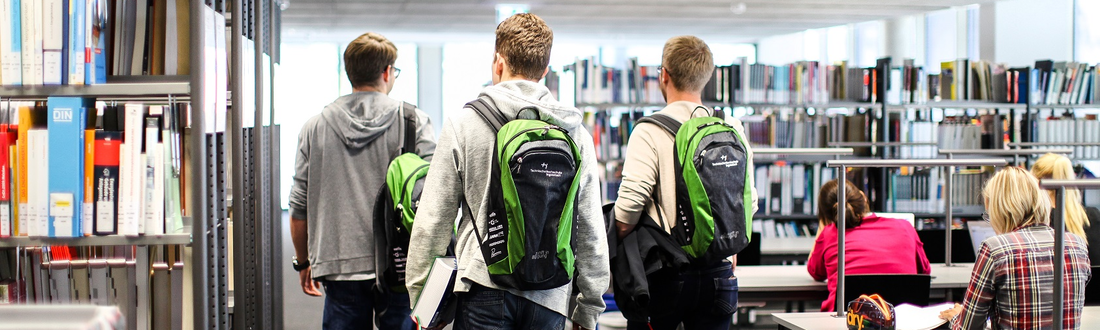 Three students with green THI backpacks walk through the library