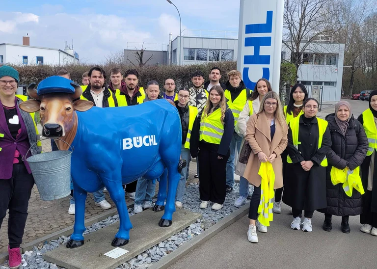 Gruppenphoto auf dem Firmengelände der Firma Büchl, im Vordergrund eine Statue einer blaugefärbten Kuh, die Teilnehmer tragen gelbe Sicherheitswesten
