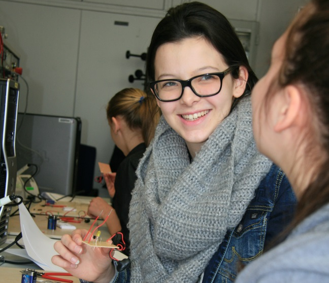 Eine Schülerin mit Brille und großem Schal unterhält sich mit einer anderen Schülerin, in der Hand hält sie ein kleines Versuchsobjekt