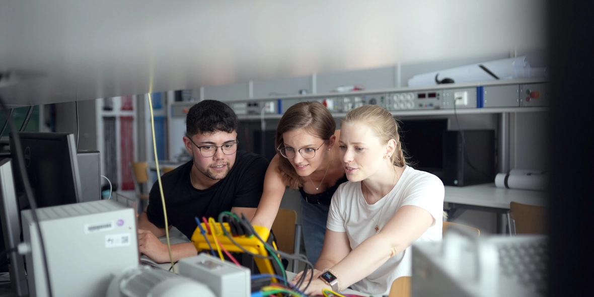 3 Studierende begutachten in einem Elektro-Fachraum elektronische Apparaturen die auf einem Tisch stehen