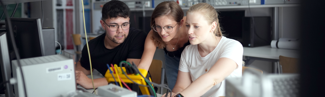 3 Studierende begutachten in einem Elektro-Fachraum elektronische Apparaturen die auf einem Tisch stehen