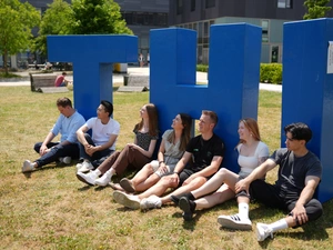 7 Studierende sitzen auf der Campuswiese und lehnen sich an Menschen-großen THI Buchstaben an