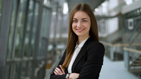 A young woman with long, loose brown hair, dressed in a white blouse and black blazer, the THI stairwell blurred in the background.