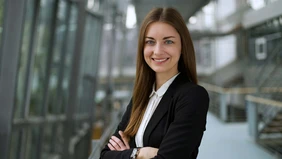 A young woman with long, loose brown hair, dressed in a white blouse and black blazer, the THI stairwell blurred in the background.