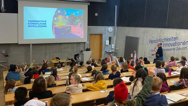 A lecture hall full of children, some raise their hands, the lecturer with microphone in hand runs to the front for the presentation. 