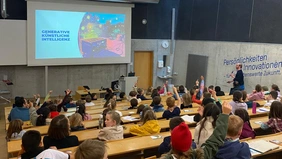 A lecture hall full of children, some raise their hands, the lecturer with microphone in hand runs to the front for the presentation. 