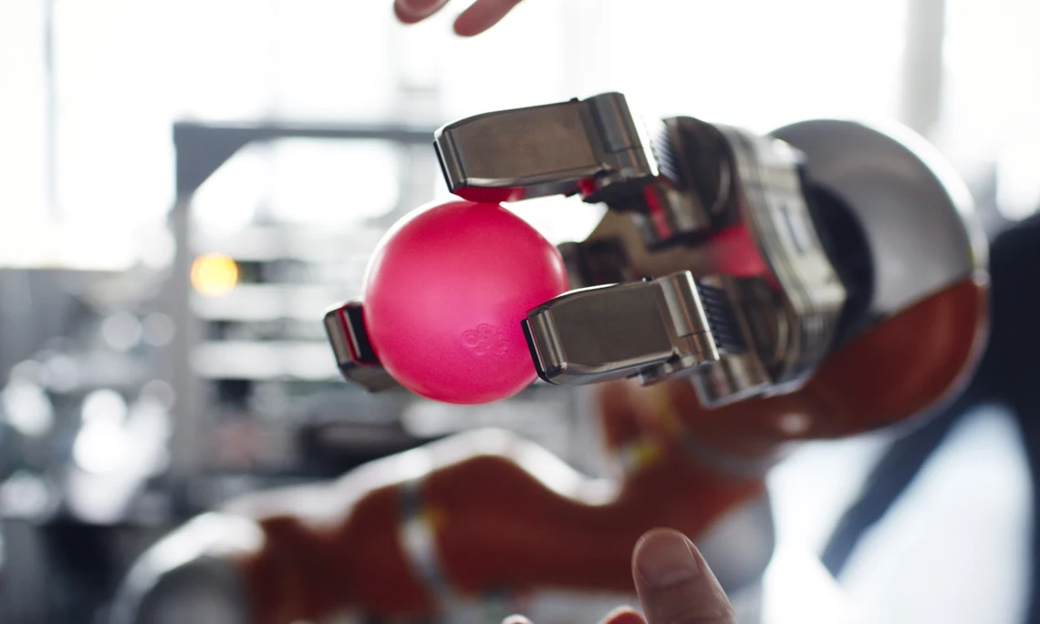 Zwei menschliche Hände und eine Robotergreifhand, die einen roten Ball hält