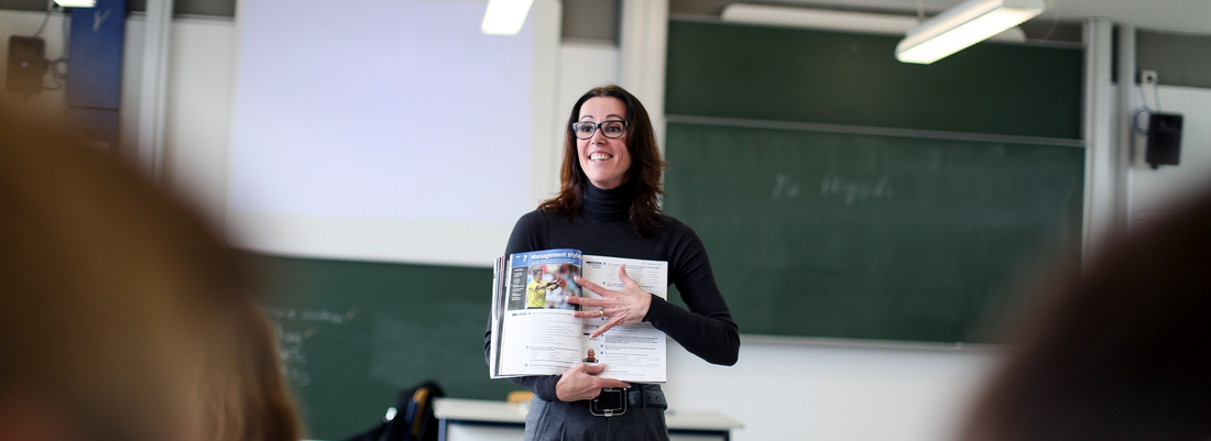 Eine Dozentin steht vor einer Tafel und erklärt Inhalte anhand eines Buches, das sie in den Händen hält