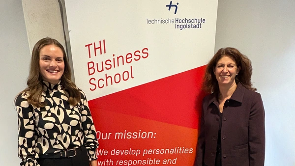 Two women stand in front of a THI Business School advertising roll-up and smile into the camera.