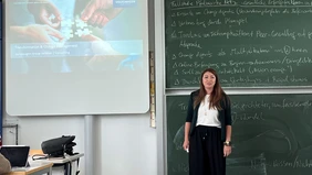 A woman standing in front of a board covered in writing, with a screen displaying a presentation to her left.