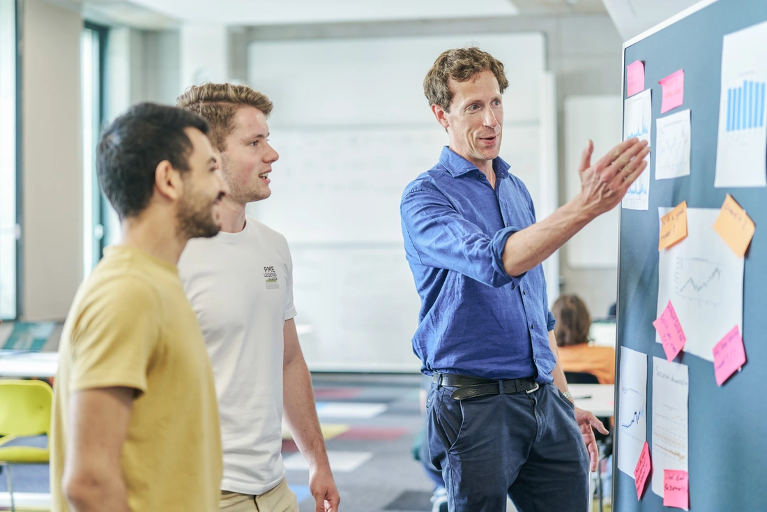 Männliche Personen vor einer Pinnwand, eine davon zeigt auf die Pinnwand