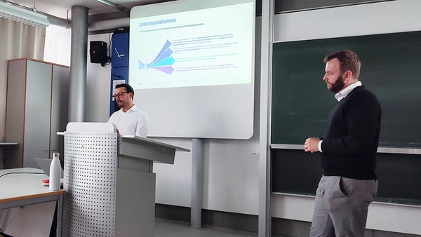 Zwei männliche Personen vor einem Whiteboard, auf dem eine Präsentation gezeigt wird.