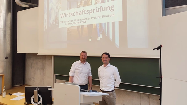 Two men smile into the camera, behind them the blackboard and screen of the lecture theatre. 