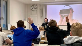 A situation in a lecture hall - from behind you see students raising their hands, in front a woman at a laptop, behind her a presentation showing a person wearing VR glasses. 