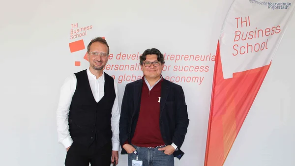 Two men stand in front of a bright wall, which is marked with the THI Business School vision: “We develop personalities with responsible and innovative mindsets and comprehensive skills in general management for success in a globalized economy".