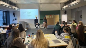 : In a seminar room, two adults are sitting on chairs at the front of the room talking to each other, while the students are sitting at the back listening to the conversation. 