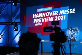 der Schatten einer Kamera ist vor einer Großbildleinwand zu sehen, die die Aufschrift "Hannover Messe Preview 2021" trägt.