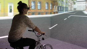 Ein Mädchen sitzt auf einem Fahrrad und trägt eine spezielle Brille. An den Wänden sieht man eine virtuelle Staße in einer Stadt.
