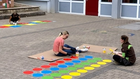 Drei Studierende bemalen mit bunten Farben einen grauen Pausenhof