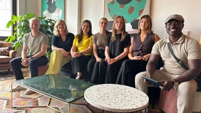 Seven people sit on a sofa and smile at the camera.