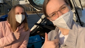 Zwei Studentinnen mit Mundschutz vor einem Auto bei dem die Kofferraumklappe geöffnet ist