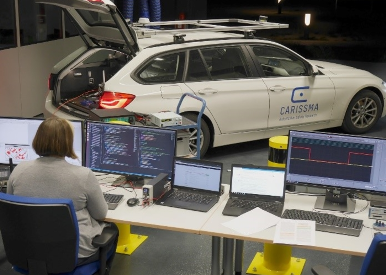 Image of a woman sitting in front of two screens. On which system tests of applications based on Car2X communication are running