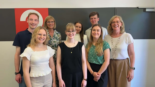 A group picture with 8 people in front of a white wall with a black stripe. 