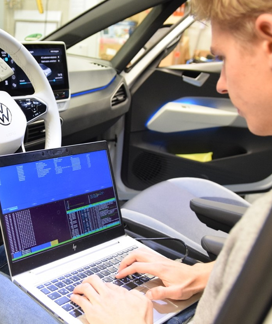 Person in Fahrzeug auf Teststand; auf dem Schoß Laptop mit Daten