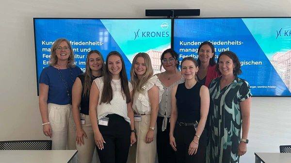 A group of eight women smiling into the camera, in the background two televisions showing the start page of the presentation.