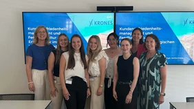 A group of eight women smiling into the camera, in the background two televisions showing the start page of the presentation.