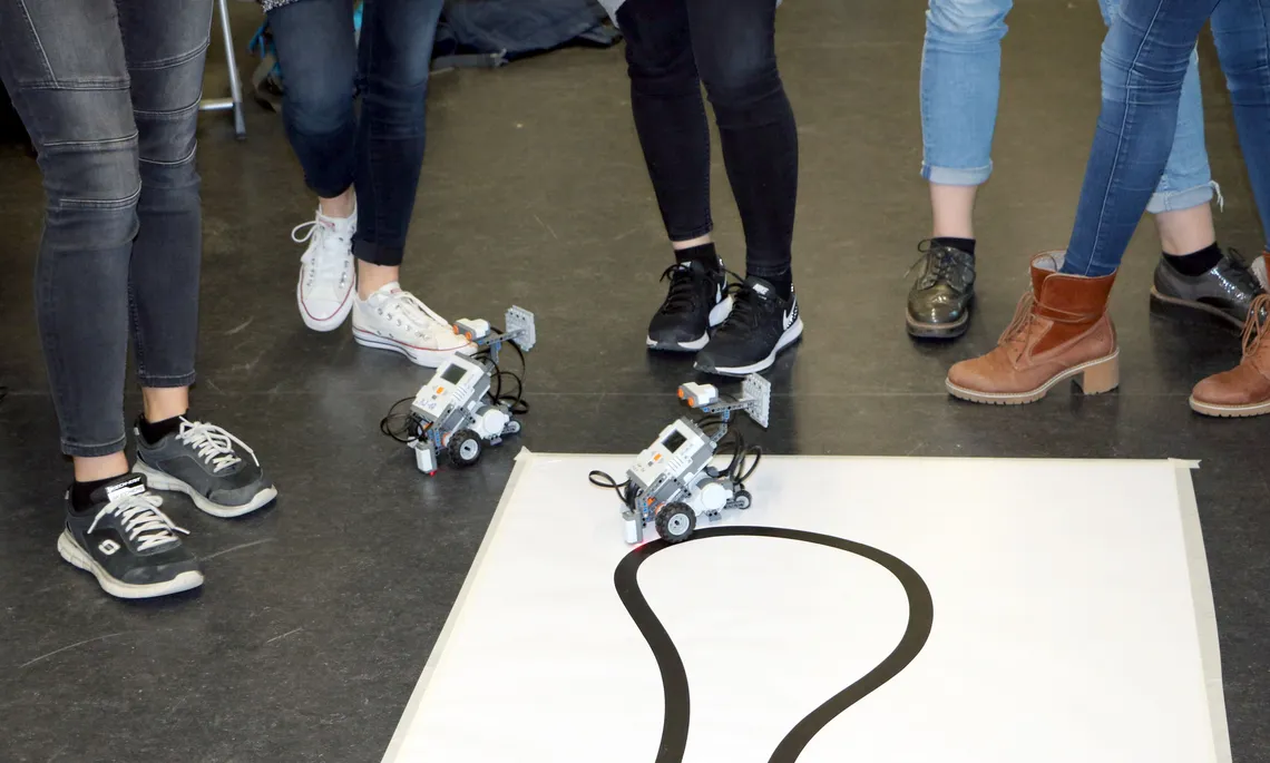 [Translate to English:] Zwei kleine Roboter fahren auf dem Boden, drumherum eine Gruppe Mädchen