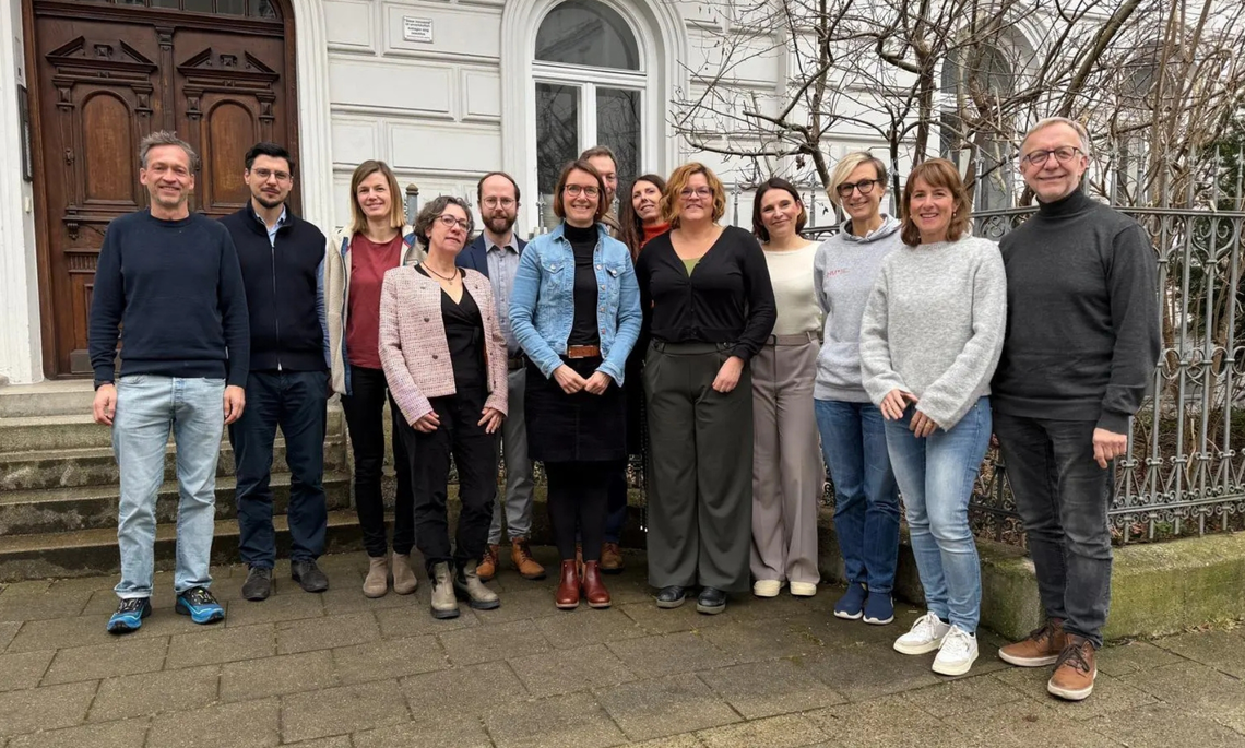 13 Personen stehen für ein Gruppenfoto vor einem Haus