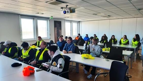 You can see a group of students sitting in a meeting room, some of them wearing yellow high-visibility vests. 