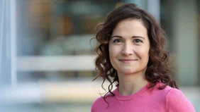 Eine Frau mit lockigen braunen Haaren und einem pinken Oberteil vor einem verschwommenen Hintergrund. 