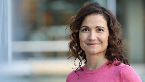 A woman with curly brown hair and a pink top against a blurred background. 