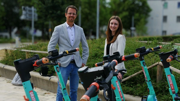 A man and a woman stand behind 5 turquoise e-scooters. 