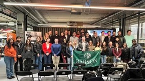 A group photo in the brigk event room, the Babson College flag is shown in the centre of the group