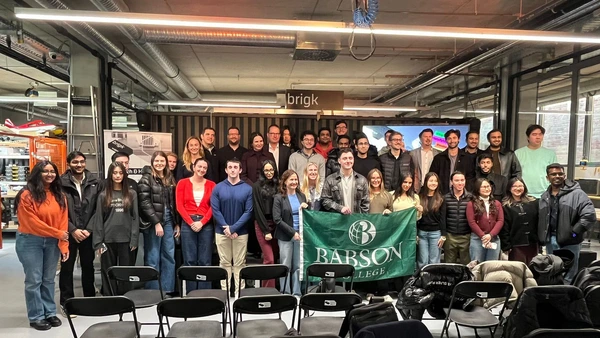 A group photo in the brigk event room, the Babson College flag is shown in the centre of the group