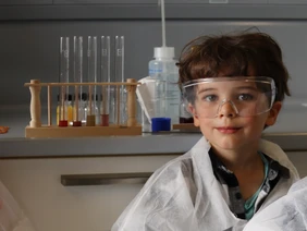 KiGa-Kind in einem Labor mit Schutzbrille und -Kleidung, im Hintergrund Reagenzgläser