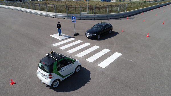 Abbildung von zwei autonom fahrender Fahrzeugen, die jeweils auf einer Seite eines Zebrastreifens stehen, den ein Dummy-Fußgänger überqueren möchte.