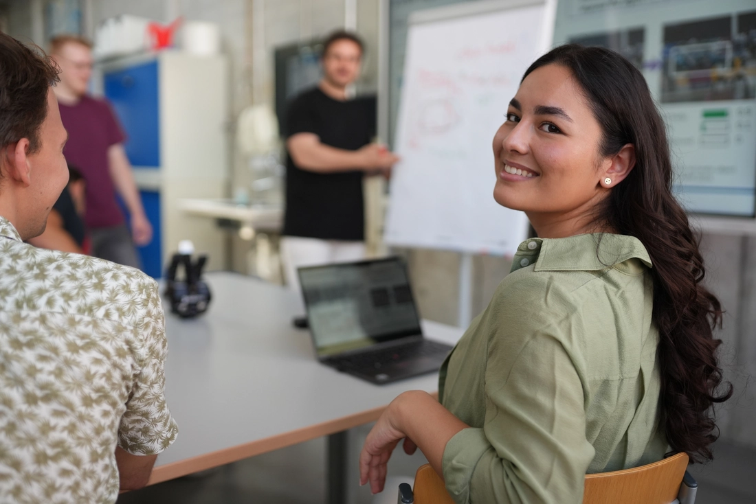 Junge Frau an Tisch, im Hintergrund Mann an Flipchart