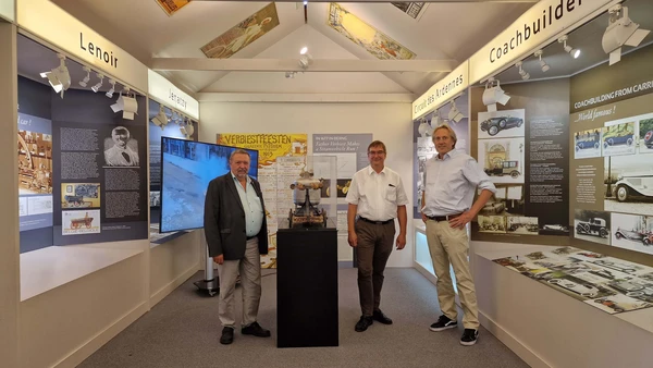Three men stand next to the replica of the first automobile in a room of the museum.