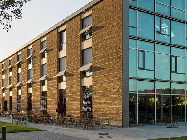 Modernes Gebäude mit Glas- und Lehmfassade, davor Sonnenschirme und Grünanlage