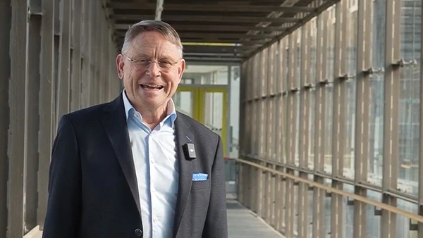 A man in a suit in a glass corridor smiles at the camera 