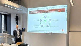 A man in a suit stands in a lecture hall, next to him you can see a presentation on the wall.