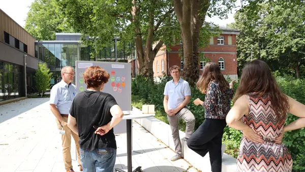 Fünf Personen im Außenbereich des Campus in Neuburg, die vor einer Pinnwand mit unterschiedlichen Tags stehen und dieskutieren