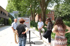 Fünf Personen im Außenbereich des Campus in Neuburg, die vor einer Pinnwand mit unterschiedlichen Tags stehen und dieskutieren