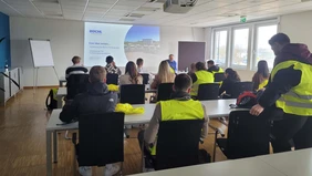 the participants of the excursion sit in a conference room and watch the presentation shown, some participants wear yellow safety vests